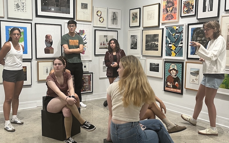 A group of students sit and stand in a gallery with artworks hung on the walls