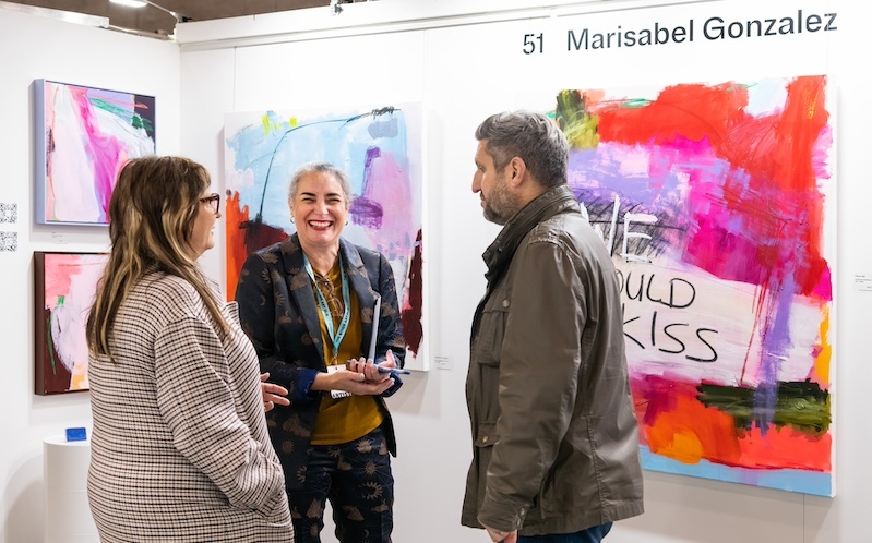 woman standing in between two people looks at camera and smiles with her abstract, vibrant, colorful artworks behind her