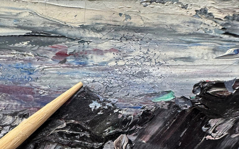 A close-up image of an oil painting with thick, textured brushstrokes in dark and muted colors. A wooden tool, possibly a conservation or restoration tool, is pointing at an area of damage where the paint has cracked and started to flake away. The cracks are concentrated in a lighter-colored section of the painting, contrasting with the surrounding darker and more intact paint. The detailed view highlights the painting’s impasto technique and the deterioration occurring in the artwork