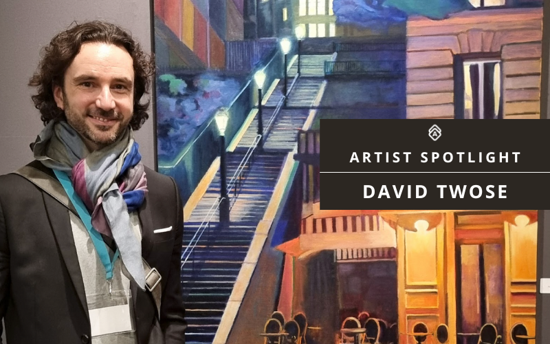 A promotional image featuring an artist spotlight for David Twose. In the foreground stands a man wearing a dark suit and a colorful patterned scarf. Behind him is a vibrant artwork depicting an urban nightscape with illuminated stairs and glowing storefronts in deep blues and warm yellows. The painting appears to show a street scene with dramatic lighting. A black overlay in the corner displays "ARTIST SPOTLIGHT" and "DAVID TWOSE" in white text, along with a geometric logo.