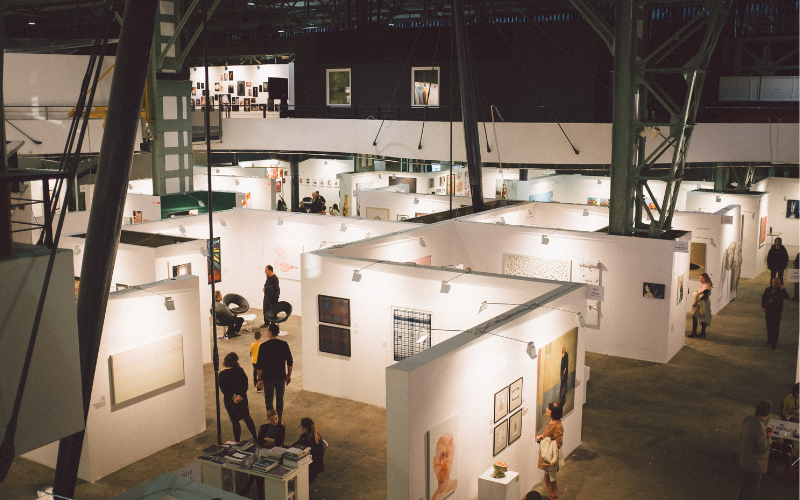 An overhead view of an indoor art fair with white partition walls creating individual display spaces. People are seen wandering and viewing the artwork, some standing and others sitting on the furniture provided. The venue has a high ceiling with industrial-style beams and hanging lights that illuminate the space
