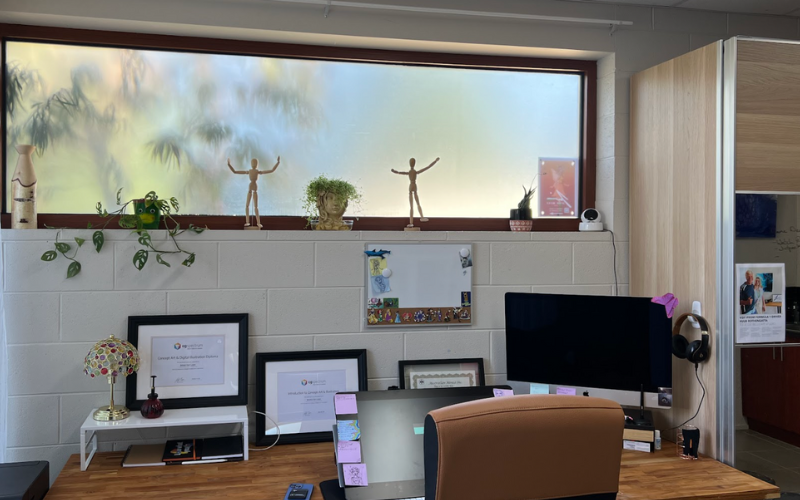 A modern and organized office space with a wooden desk, two computer monitors, and framed certificates. The desk has sticky notes attached to the monitor, a small stained glass lamp, and a few office supplies. A brown leather office chair sits in front of the desk. The white brick wall behind the desk features a whiteboard with a few pinned items. Above the wall, a long frosted window allows natural light to enter, with decorative elements on the windowsill, including two wooden artist mannequins, a small potted plant in a head-shaped planter, a cascading plant in a green pot, a vase, and a framed poster.