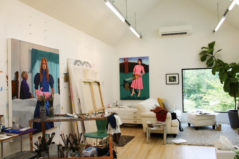 This image shows a bright and spacious art studio that belongs to Sharon Shapiro. It features high ceilings, large windows, and a cozy seating area. Bold, colorful paintings, including a woman in pink with a chainsaw, decorate the space. A cluttered artist's table with paintbrushes and tools is pictured.